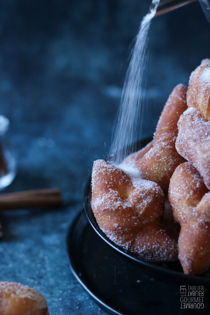Gros plan sur une bugne que l'on saupoudre généreusement de sucre en poudre