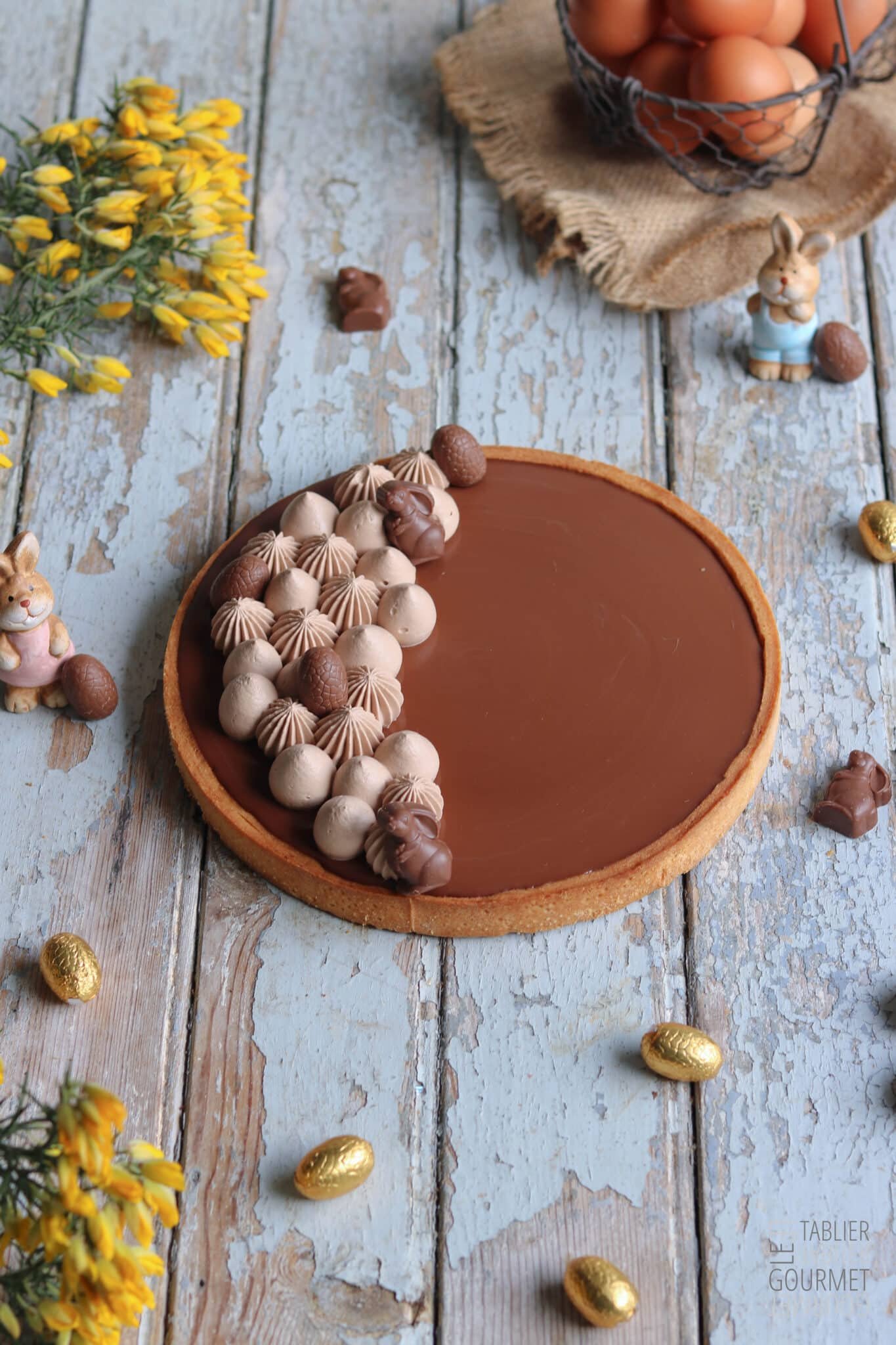 Une tarte au chocolat et caramel avec de la ganache montée caramélisée pochée dessus et sur laquelle sont déposées des petits sujets de Pâques en chocolat